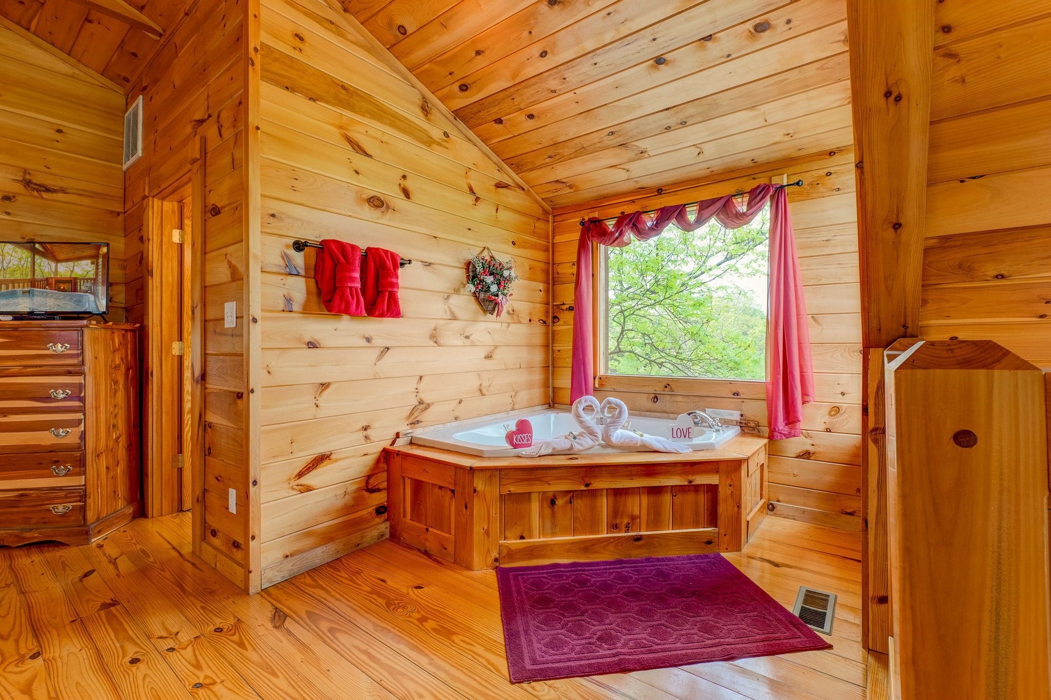 Ein Holzblockhaus-Interieur mit Holzboden, einer Holzwanne, einem Fenster mit rosa Vorhängen und einem roten Handtuch, das an der Wand hängt.