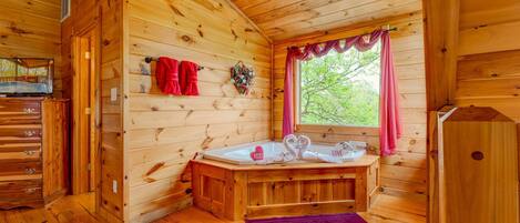 A wooden cabin interior with a wooden floor, a wooden bathtub, a window with pink curtains, and a red towel hanging on the wall.