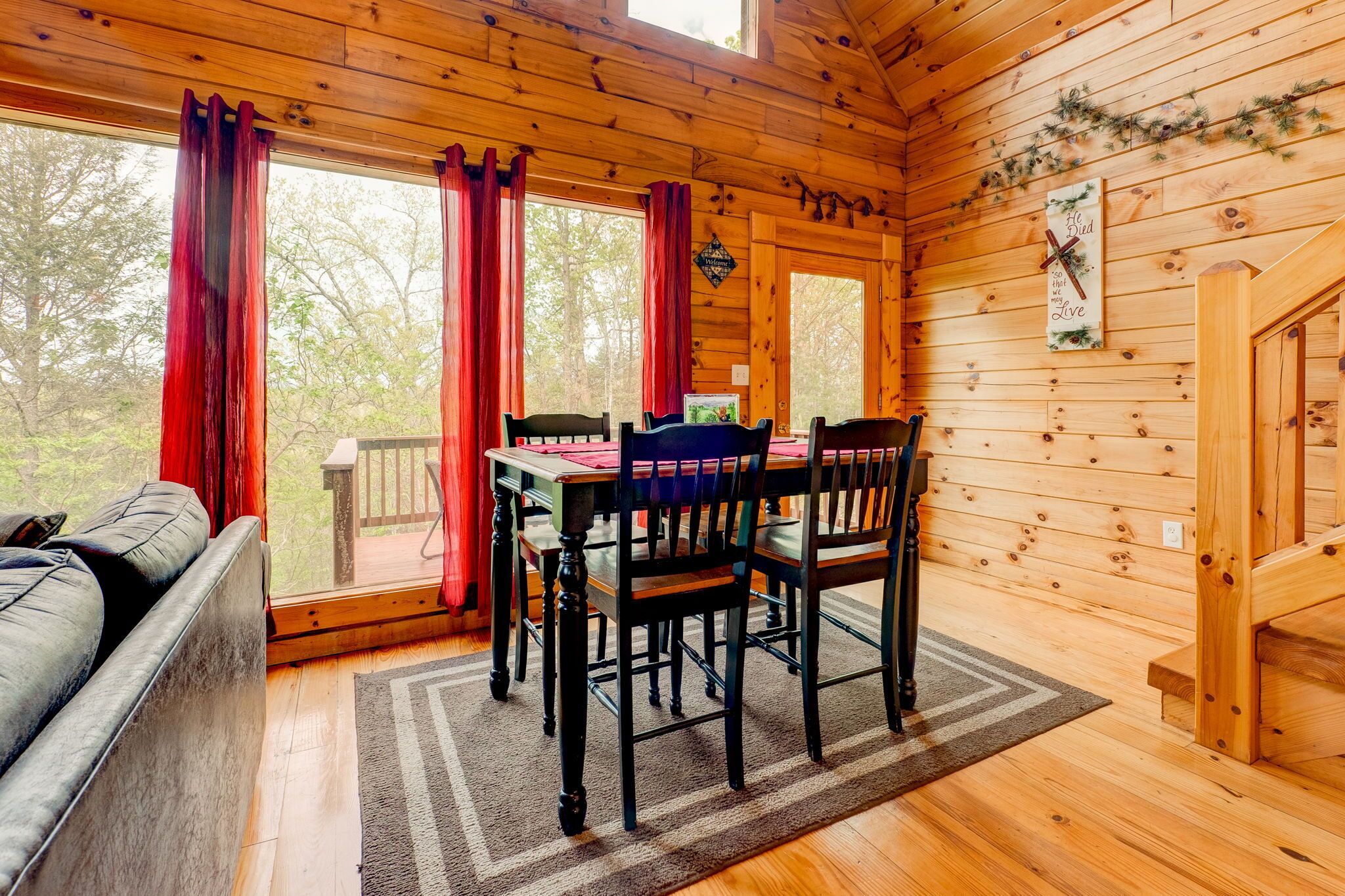 A wooden cabin interior with a dining table and chairs, a sofa, and large windows with red curtains.