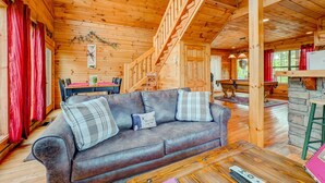 A cozy cabin interior with a wooden staircase, a sofa with plaid cushions, a dining area, and a stone fireplace.