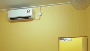 Een kleine kamer met gele wanden, een bed, een ventilator, een airconditioner en een zichtbare badkamers deur.