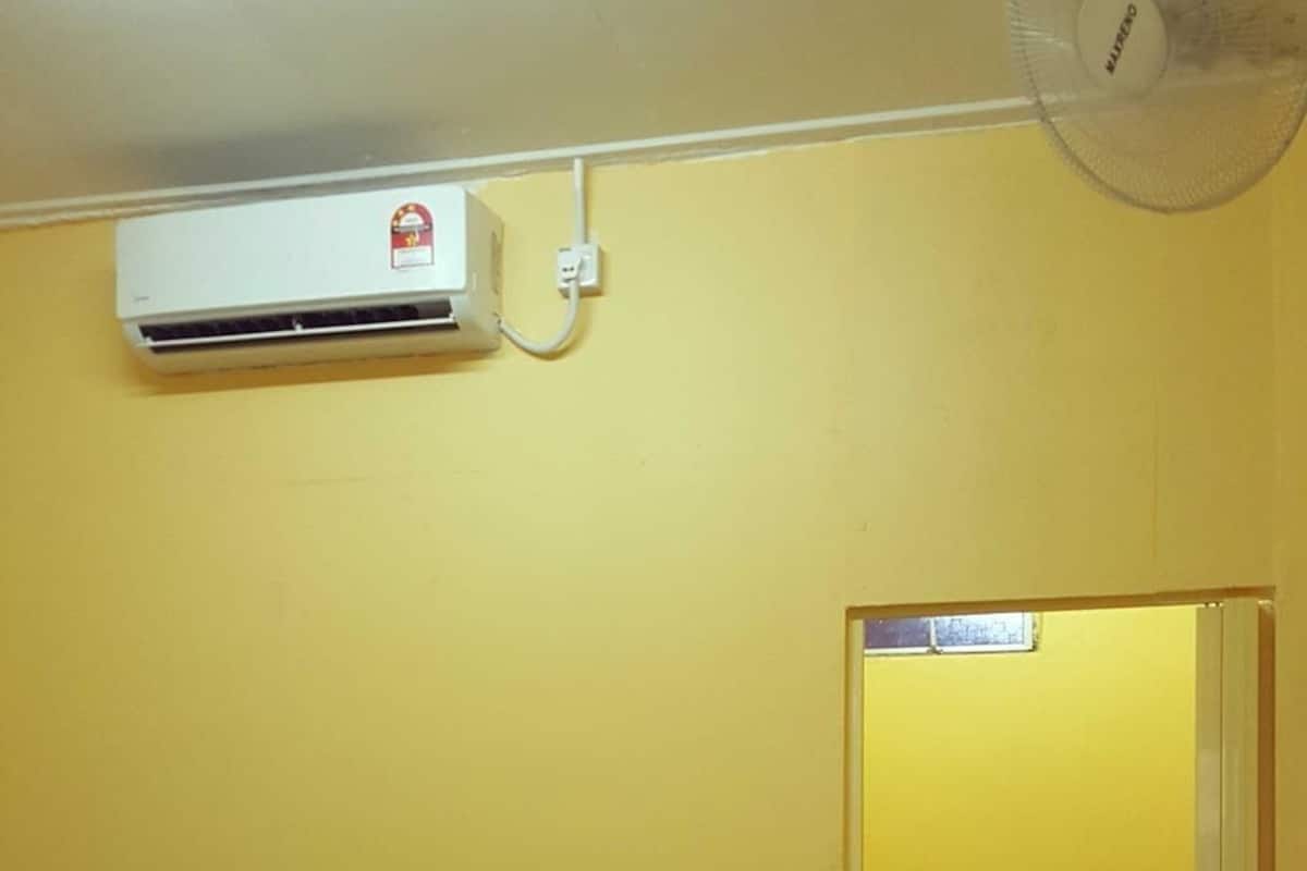 Een kleine kamer met gele wanden, een bed, een ventilator, een airconditioner en een zichtbare badkamers deur.