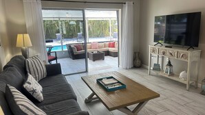 A living room with a sofa, coffee table, TV, and a view of an outdoor pool area. -  Paradise Cove -- A perfect Beach Getaway (Bradenton)
