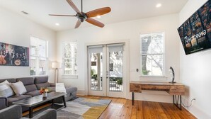 Un salon moderne avec un canapé, une table basse et un bureau avec une lampe. De grandes fenêtres sont équipées de stores, et on y trouve également un ventilateur de plafond ainsi qu’un téléviseur fixé au mur.