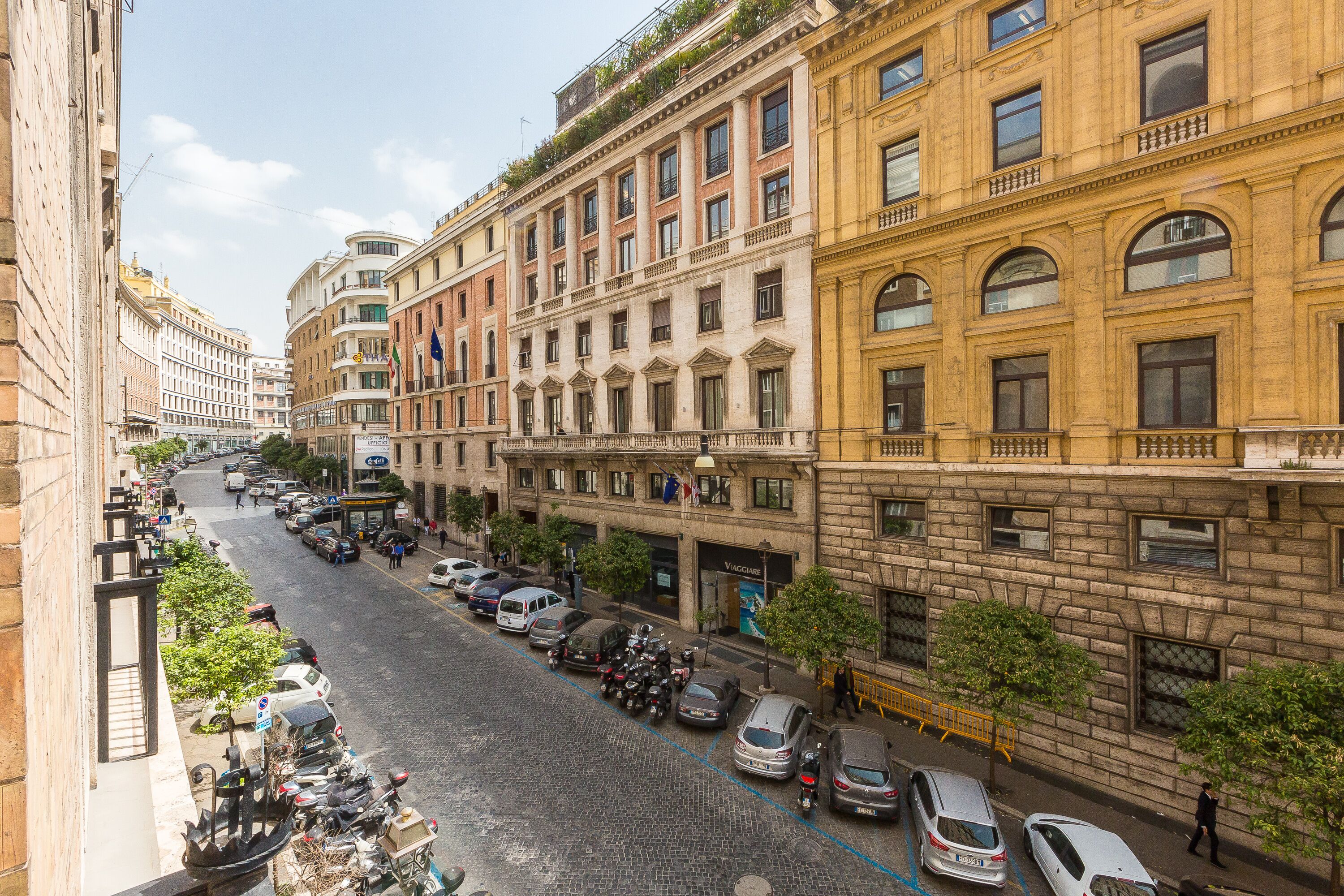 Une vue d’une rue d’une ville avec des bâtiments historiques, des voitures garées et un ciel dégagé.