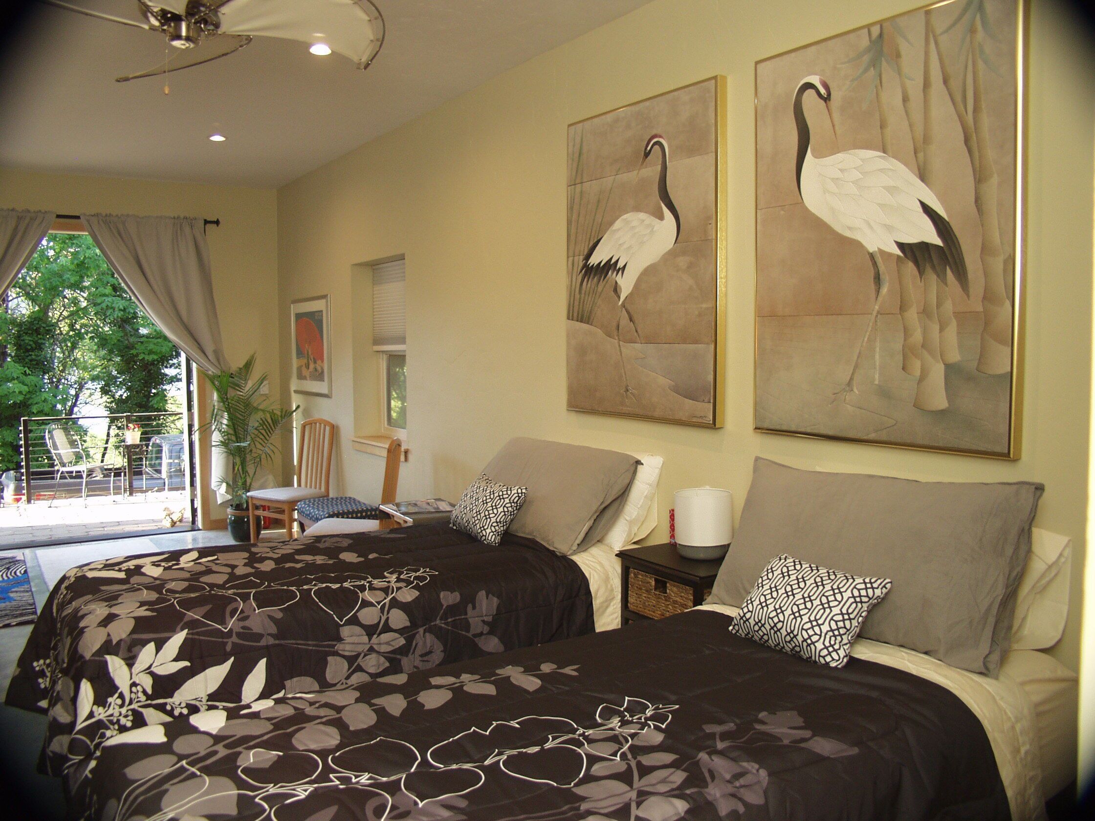 A hotel room with two beds, a ceiling fan, a window with curtains, a potted plant, and artwork on the walls.