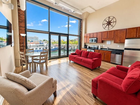 A spacious living area with a large window, a red sofa, a striped armchair, and a kitchen with modern appliances.