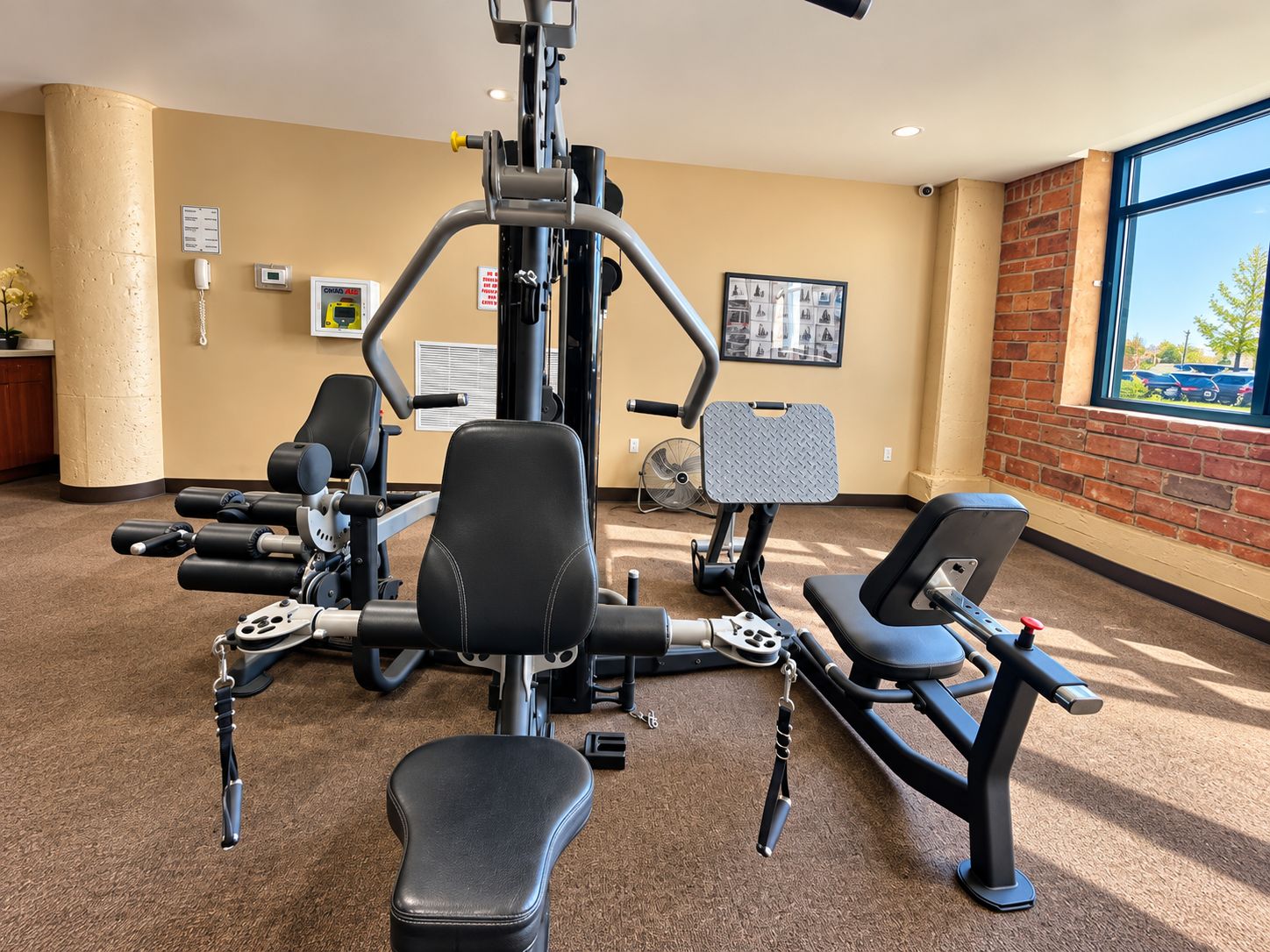 A gym with various exercise machines, a wall-mounted fan, and a window with a view of the outside.