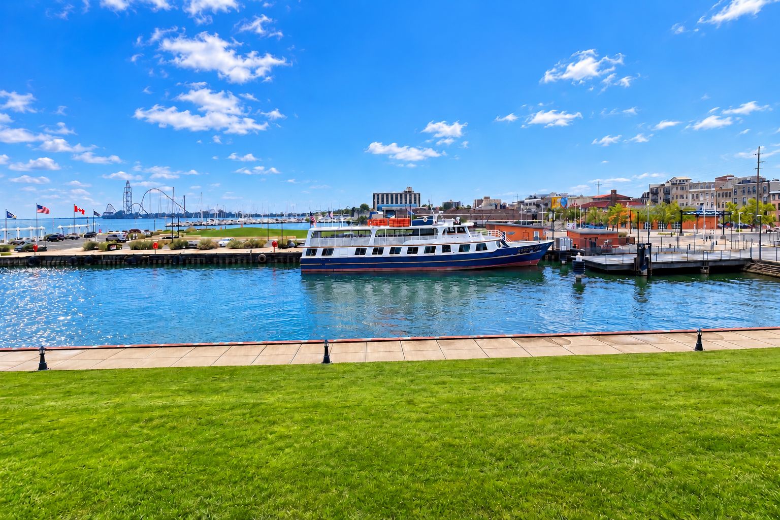 A waterfront area with a large boat docked at a marina, surrounded by a grassy promenade and a cityscape in the background.