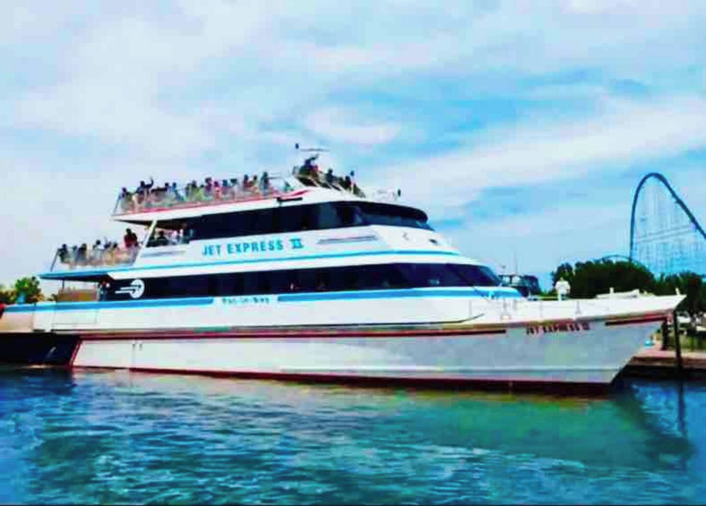 Un grand yacht blanc, sur lequel on pouvait lire les mots « JET EXPRESS II », était amarré à un quai, avec des montagnes russes en arrière-plan.