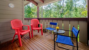 Une terrasse couverte avec un plancher en bois, des fauteuils Adirondack rouges et bleus, une petite table ronde et une horloge au mur.