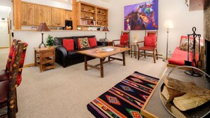 Un salon chaleureux avec un canapé, des fauteuils, une table basse et une cheminée. On y trouve une bibliothèque remplie de livres et d’objets décoratifs, ainsi qu’un tableau accroché au mur.