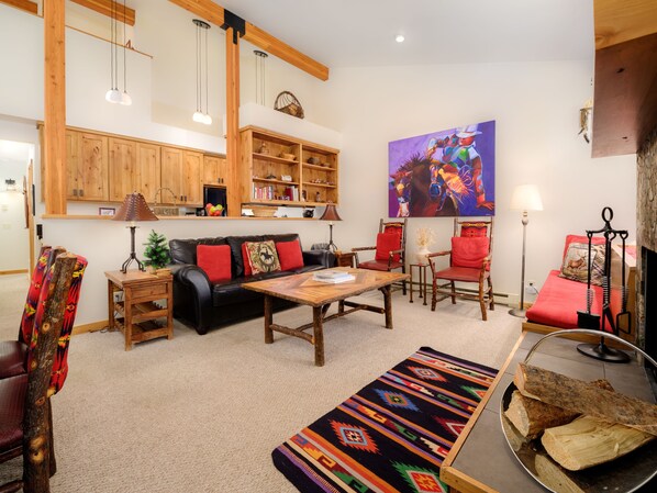Un salon chaleureux avec un canapé, des fauteuils, une table basse et une cheminée. On y trouve une bibliothèque remplie de livres et d’objets décoratifs, ainsi qu’un tableau accroché au mur.
