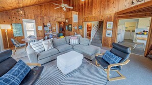 A cozy cabin interior with a large gray sectional sofa, two black leather chairs, a white ottoman, and a patterned rug. There are ceiling fans, wooden walls, and a staircase leading to a kitchen area. - Hatteras Island Charmer! (Salvo)