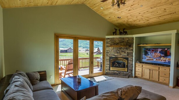A cozy living room with a stone fireplace, a television, and a large window overlooking a deck. - Family Friendly - On Ten Acres 3 Miles to Town - Stunning Views and Country Feel (Crested Butte)