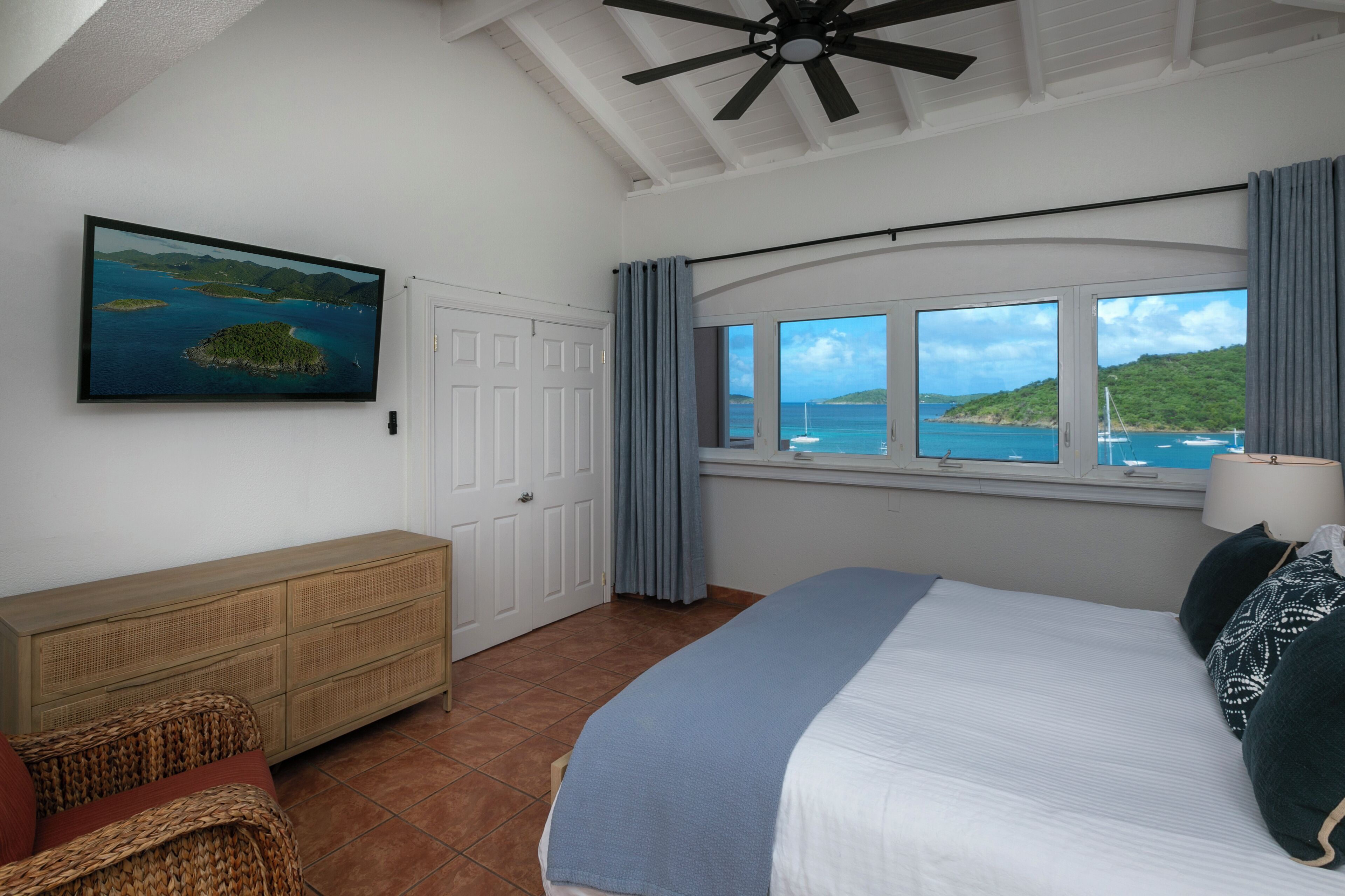 A bedroom with a bed, a wooden dresser, a window with a view of the sea, and a ceiling fan.