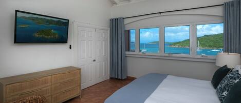 A bedroom with a bed, a wooden dresser, a window with a view of the sea, and a ceiling fan.
