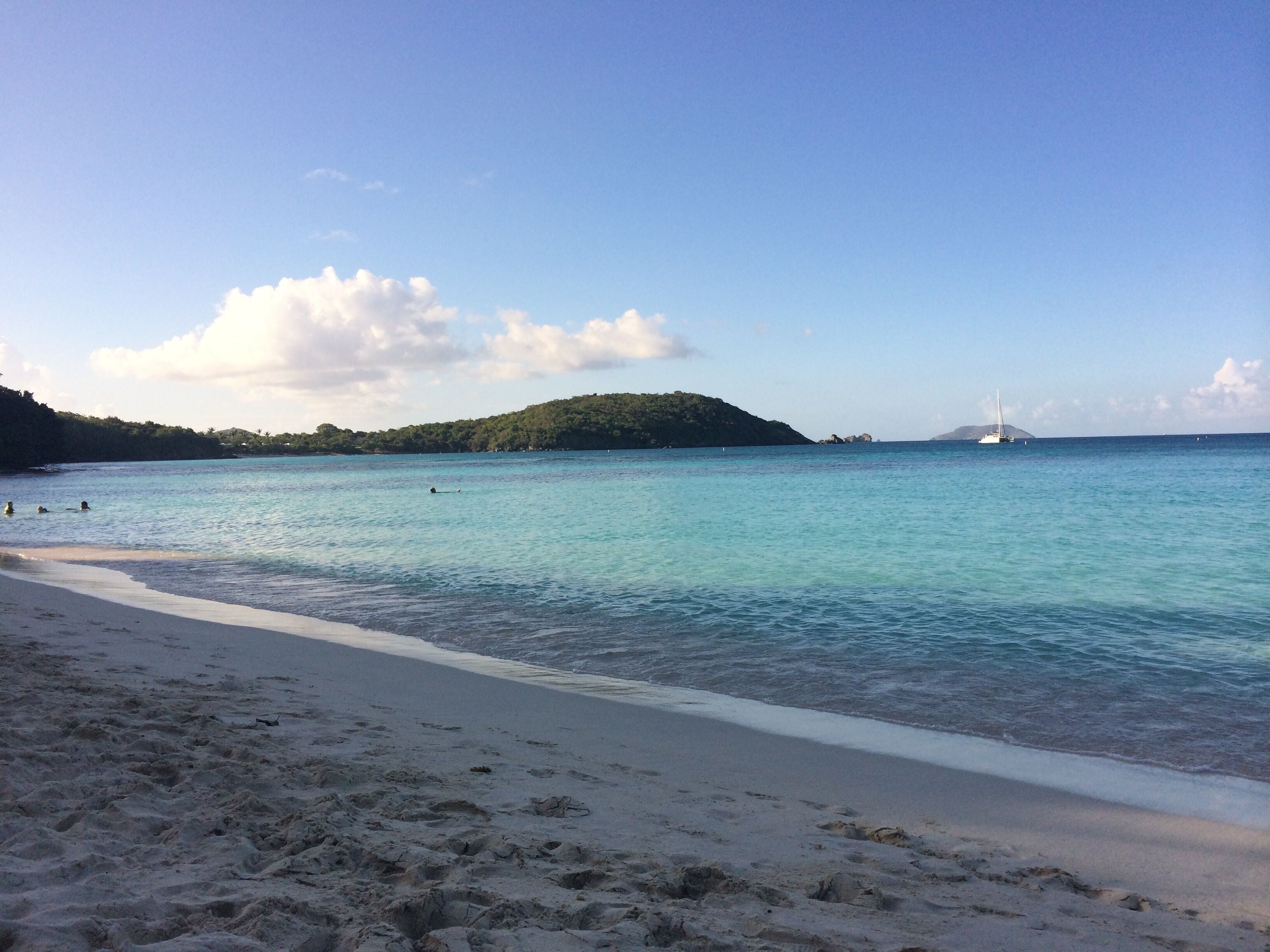 A beach with clear turquoise water, a sandy shore, and a distant sailboat.