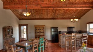 Cozinha rústica com vigas de madeira, mesa de jantar, cadeiras, geladeira e bancada com cadeiras.