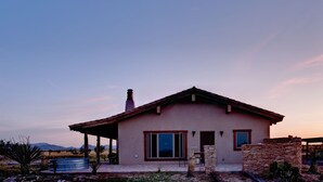 Uma casa térrea com telhado plano, chaminé e uma grande janela. Há uma área de pátio com mesa e cadeiras, e uma piscina pequena.