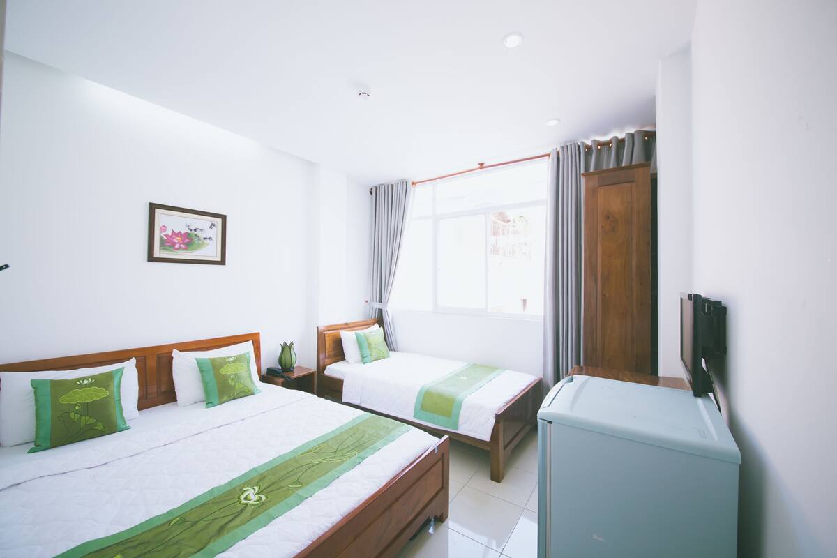 Quarto de hotel com duas camas, cabeceira de madeira, mesinha de cabeceira com um vaso, janela com cortinas e uma televisão sobre um suporte.