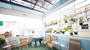 Une terrasse ensoleillée avec une table ronde et des chaises en osier, entourée de plantes en pot et dotée d’un sol carrelé.