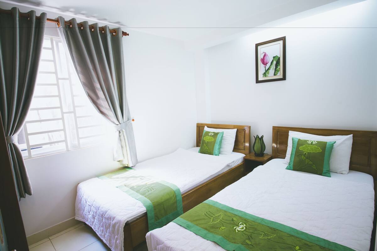 Quarto de hotel com duas camas de solteiro, cada uma com colcha e almofadas verdes e brancas. Há uma moldura com uma imagem de flor na parede, e uma janela com cortinas.