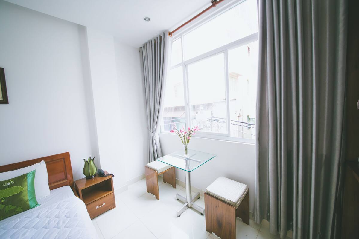Quarto de hotel com uma cama, uma mesinha com um vaso de flores, uma mesa de cabeceira de madeira e uma janela com cortinas.