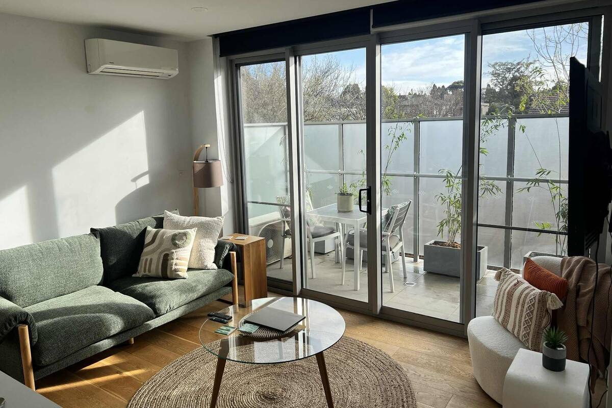Ein modernes Wohnzimmer mit einem grünen Sofa, einem Glastisch und einem weißen Sessel. Eine große Schiebetür aus Glas führt zu einem Außenbereich mit Tisch und Stühlen.