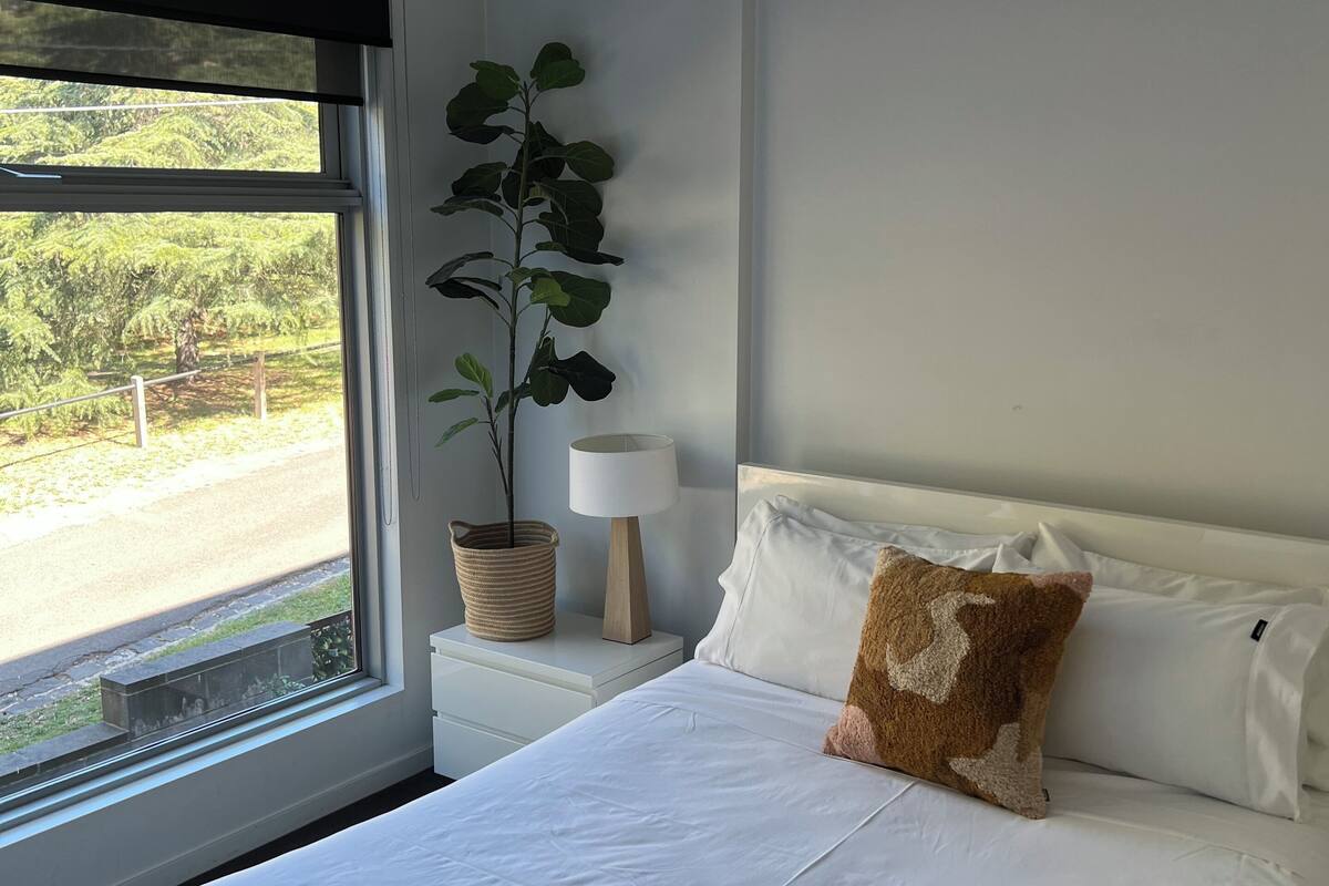 Ein Schlafzimmer mit Bett, Nachttisch, Lampe, Topfpflanze und Fenster mit Blick ins Grüne.