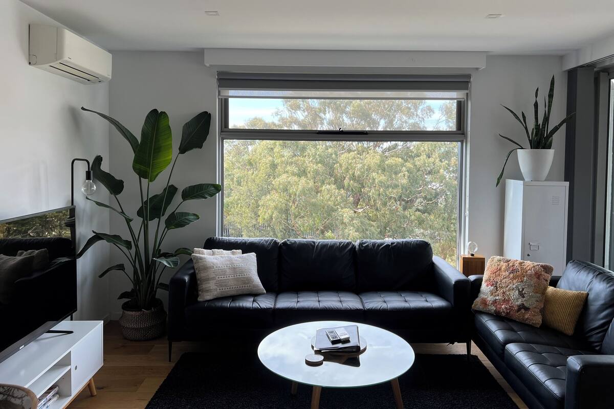 Ein modernes Wohnzimmer mit einer schwarzen Ledersofa, einem weißen Couchtisch und einem großen Fenster mit Blick auf Bäume.