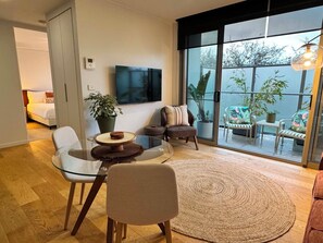 A modern living room with a glass table, comfortable seating, a large window with a view of the outdoors, and a flat-screen TV mounted on the wall. - Centrepoint Units Heidelberg (Heidelberg)