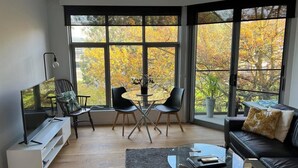 A modern living room with a TV, seating, and large windows offering a view of autumn foliage. - Centrepoint Units Heidelberg (Heidelberg)