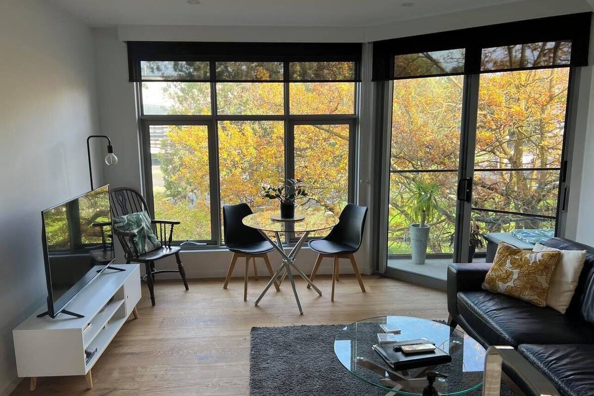 Ein modernes Wohnzimmer mit Fernseher, Sitzgelegenheiten und großen Fenstern mit Blick auf Herbstlaub.