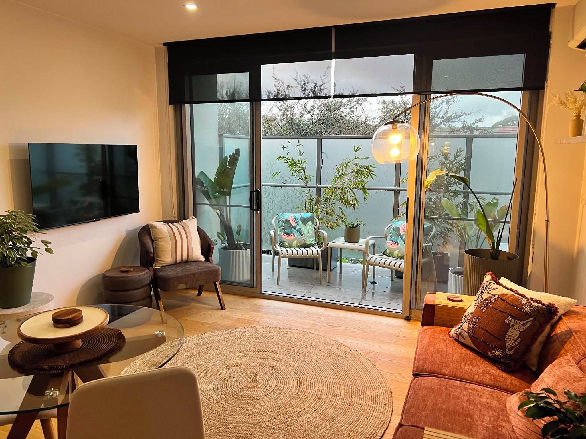 Una sala de estar moderna con un amplio ventanal corredero que da a un balcón con plantas y vistas a árboles.