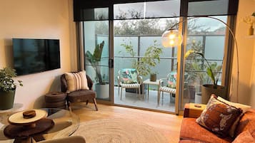 Una sala de estar moderna con un amplio ventanal corredero que da a un balcón con plantas y vistas a árboles.