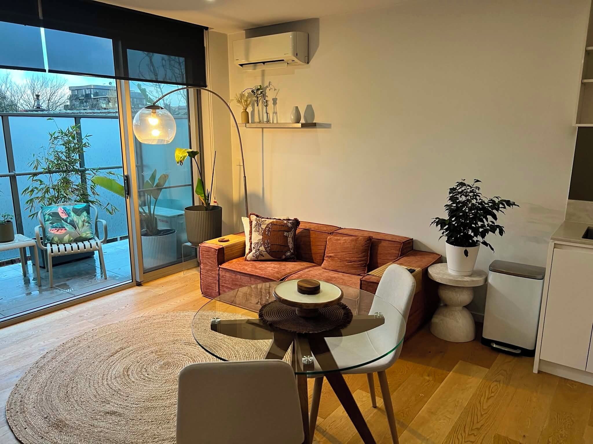 A modern living room with a glass table, a sofa with cushions, a floor lamp, and a potted plant.