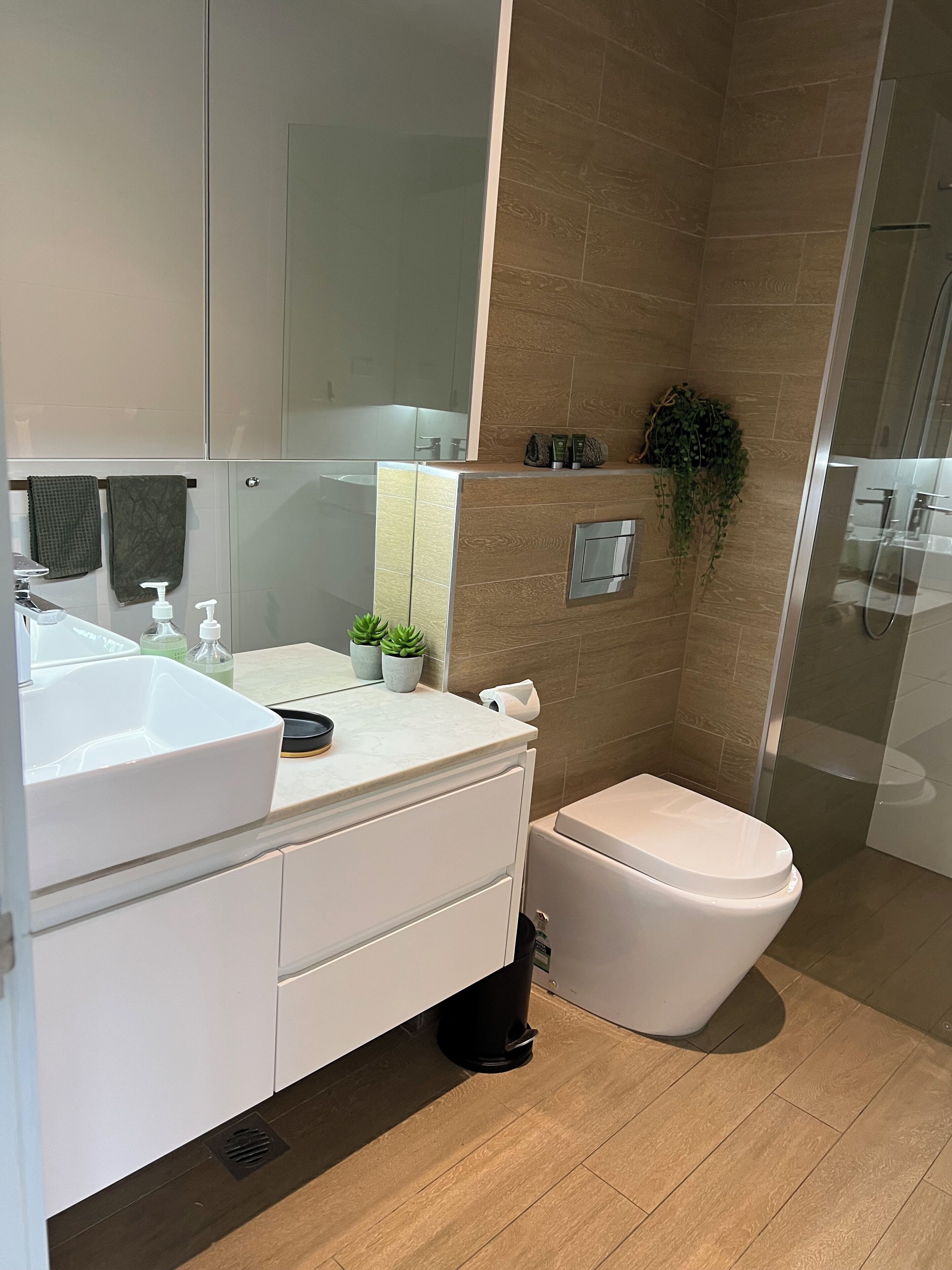A modern bathroom with a white sink, mirror, and toilet. There is a small potted plant on the counter and a shower area with glass doors.