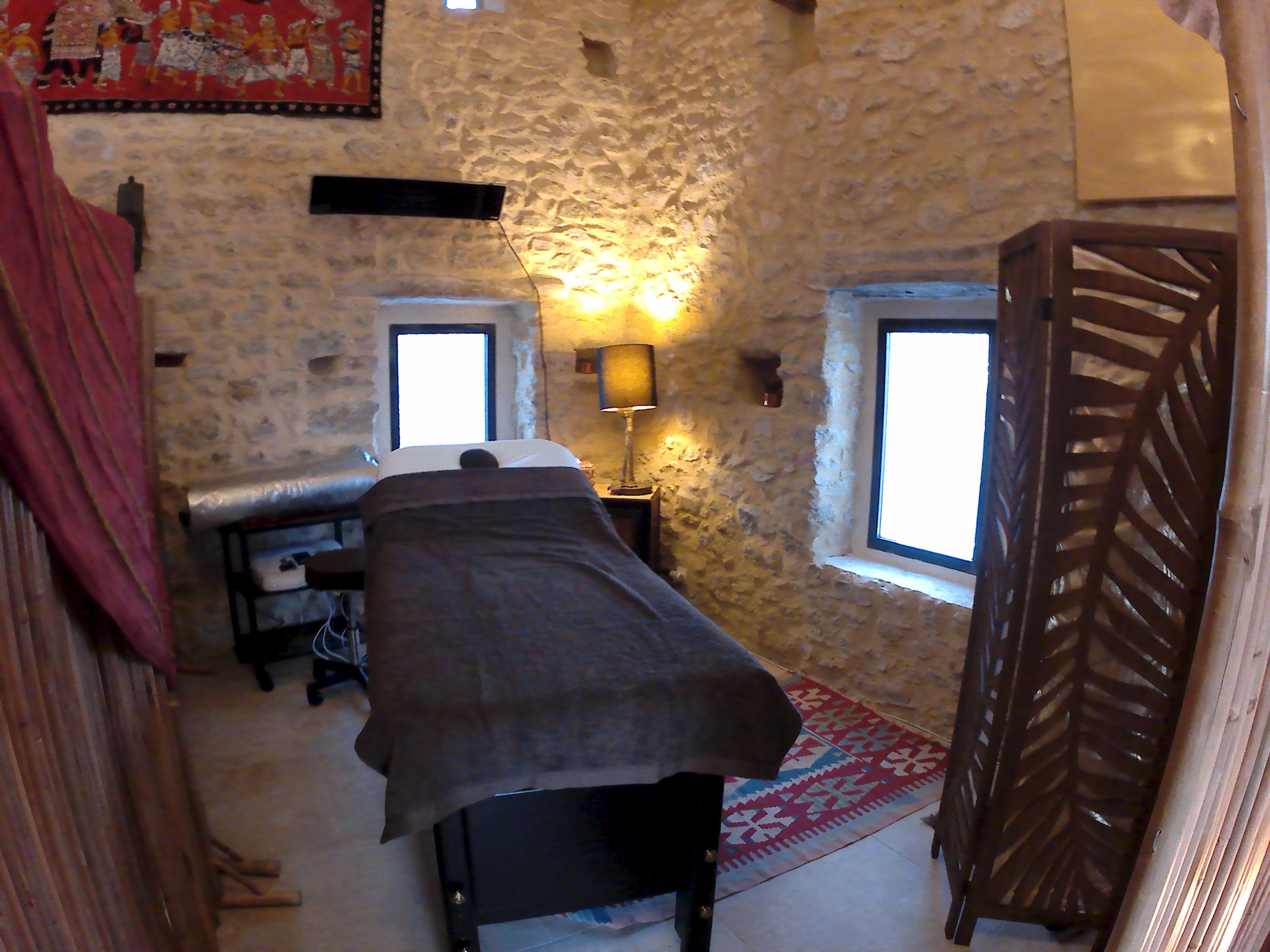 A room with a stone wall, a bed, a window, a lamp, and a patterned rug.