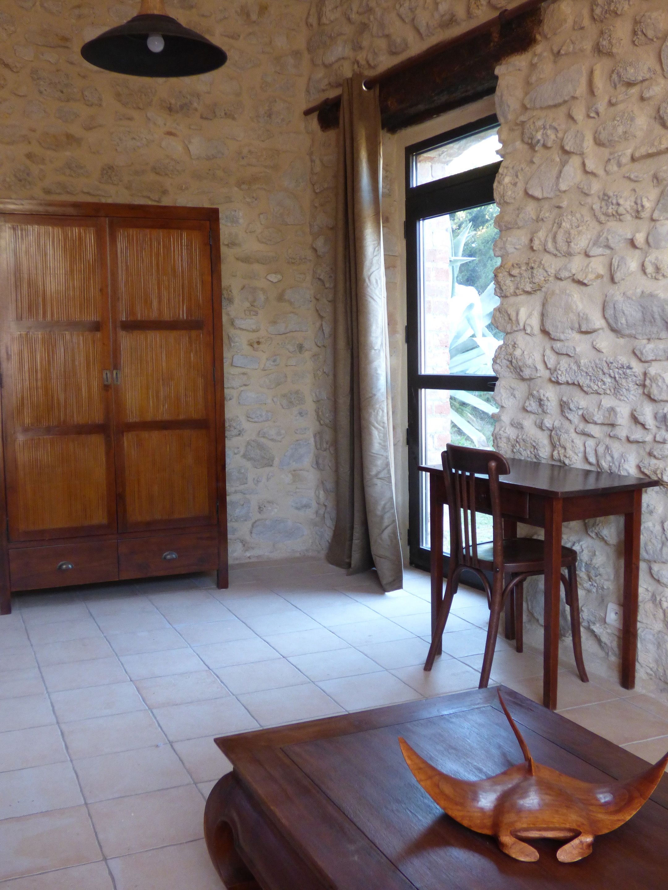 Una habitación con pared de piedra, un armario de madera, una mesa de comedor con sillas y una ventana con cortinas.
