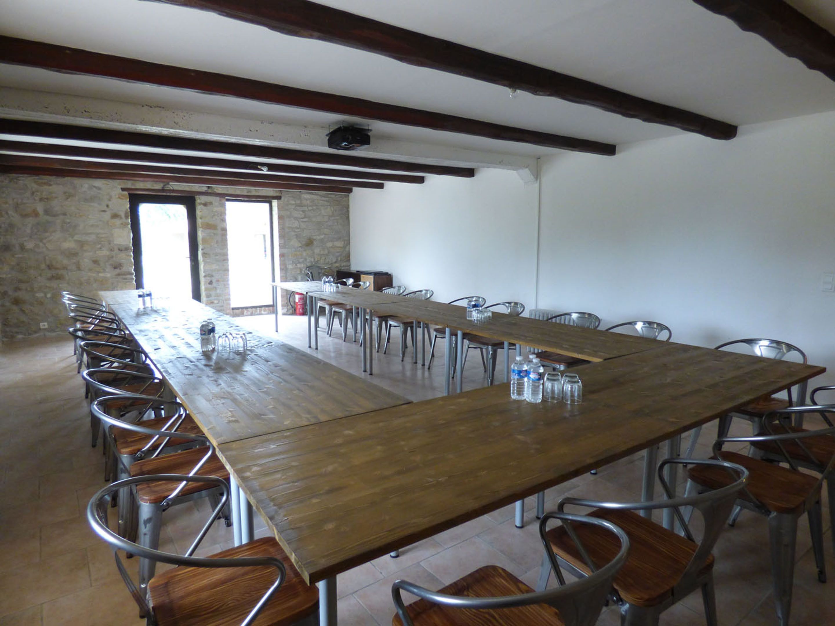 Une longue table à manger entourée de chaises en métal, un mur en pierre et des poutres en bois au plafond.