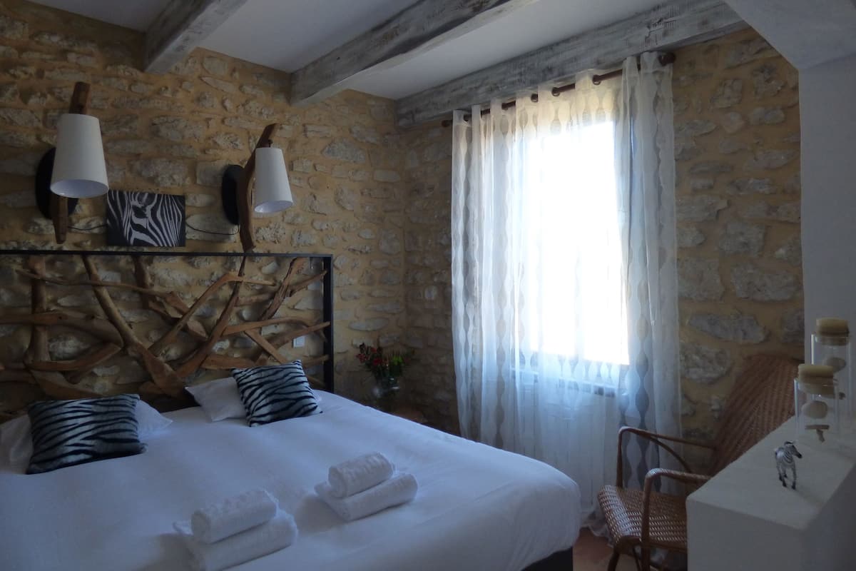Een slaapkamer met een stenen muur, een bed met witte lakens, twee lampen, een kussen met luipaardprint en een stoel.