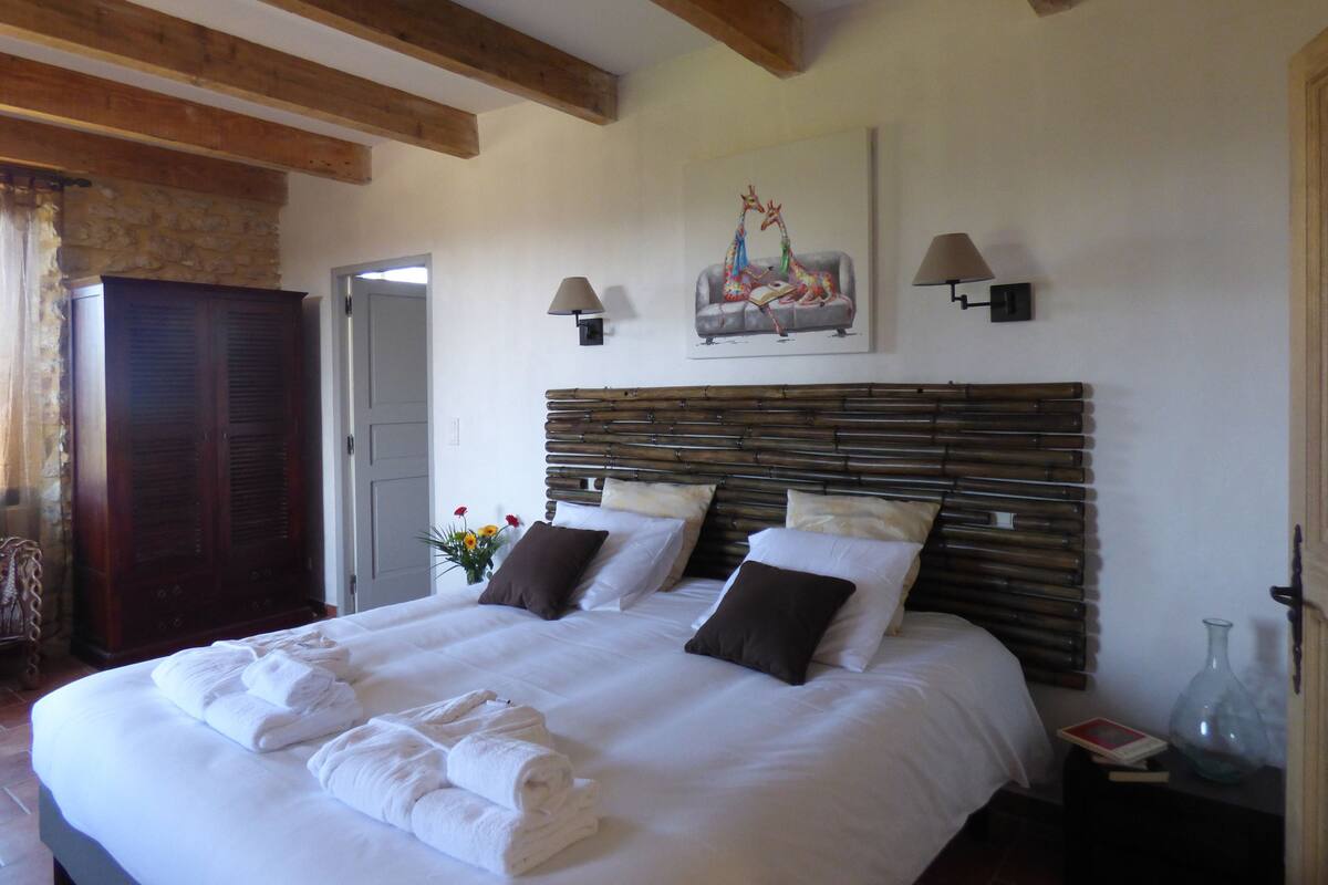 Een net opgemaakt bed met witte lakens en handdoeken, een houten hoofdbord en een schilderij van giraffen aan de muur.