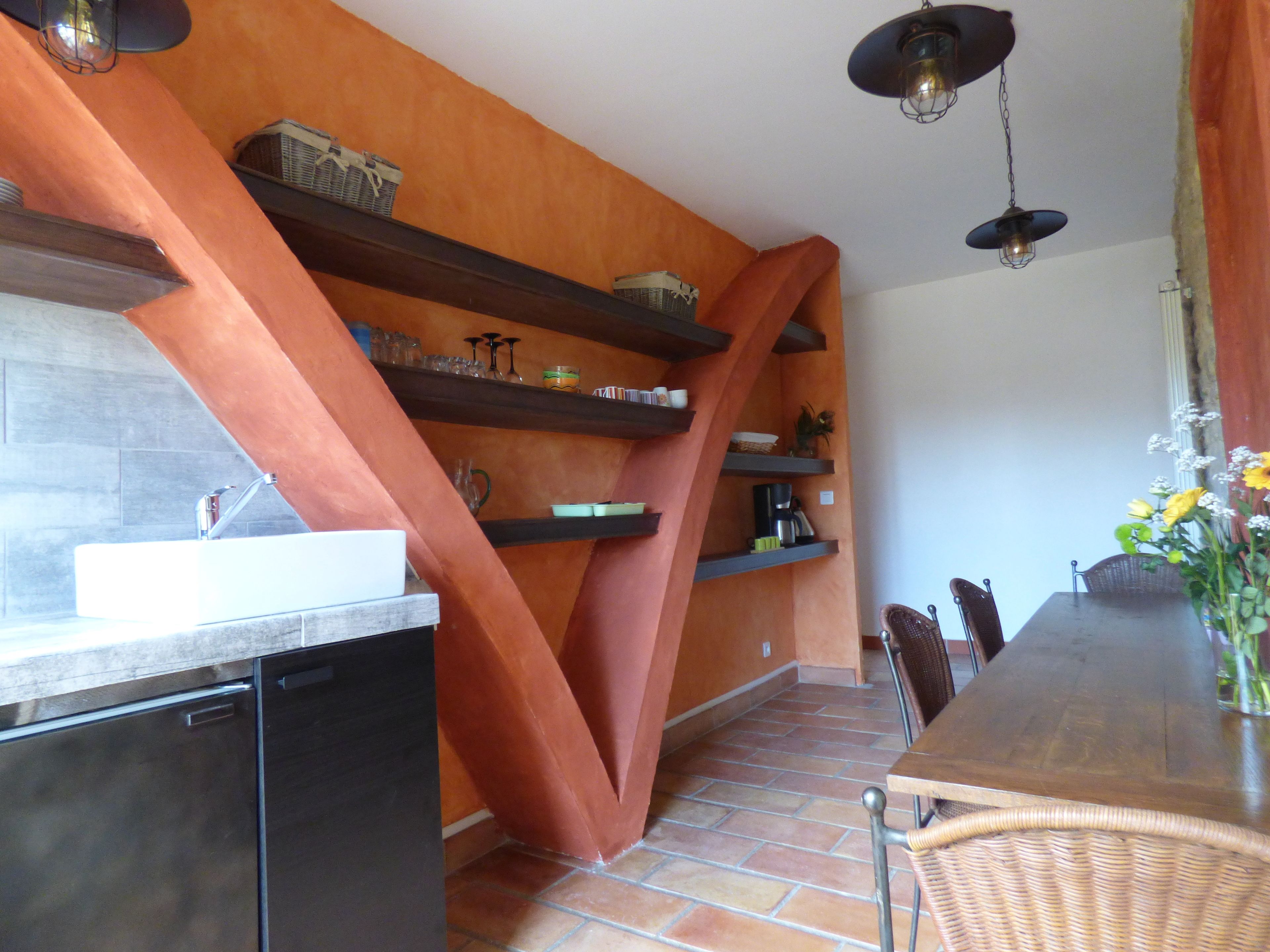 A kitchen with a dishwasher, a sink, and a dining area with a table and chairs. There are shelves with various items and a hanging light fixture.