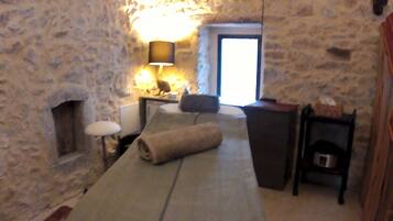 A stone-walled room with a bed, a lamp, a small table, and a window.