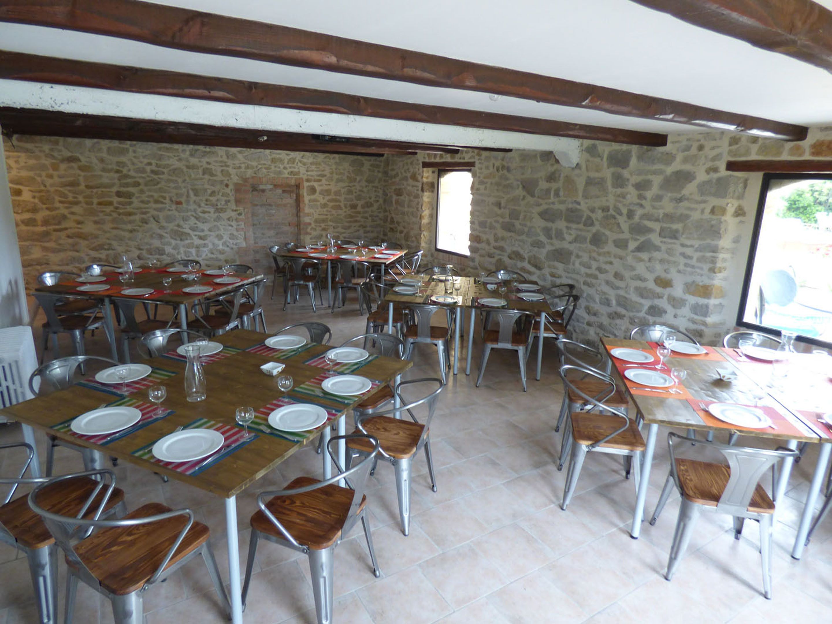 Uma sala de jantar com paredes de pedra e vigas de madeira, com várias mesas postas com pratos e copos.