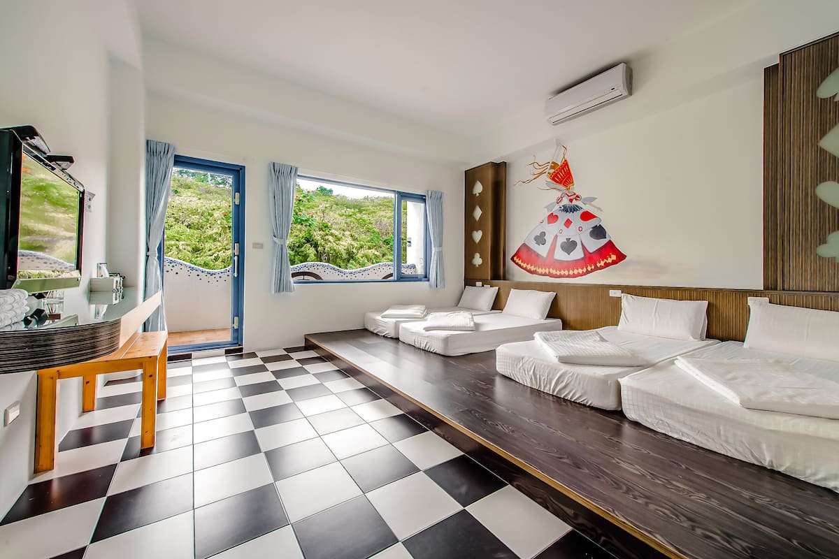 A hotel room with a checkered floor, a wooden bench, a bed with white bedding, a desk with a mirror, and a view of greenery through the window.