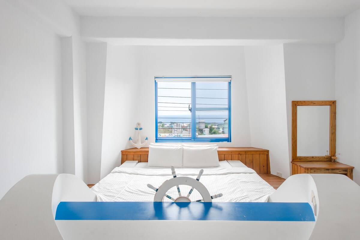 A bedroom with a bed, a wooden nightstand, a mirror, and a window with a view of buildings.
