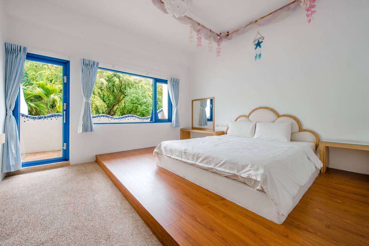A bedroom with a wooden floor, a large bed, a nightstand, a window with curtains, and a view of greenery outside.