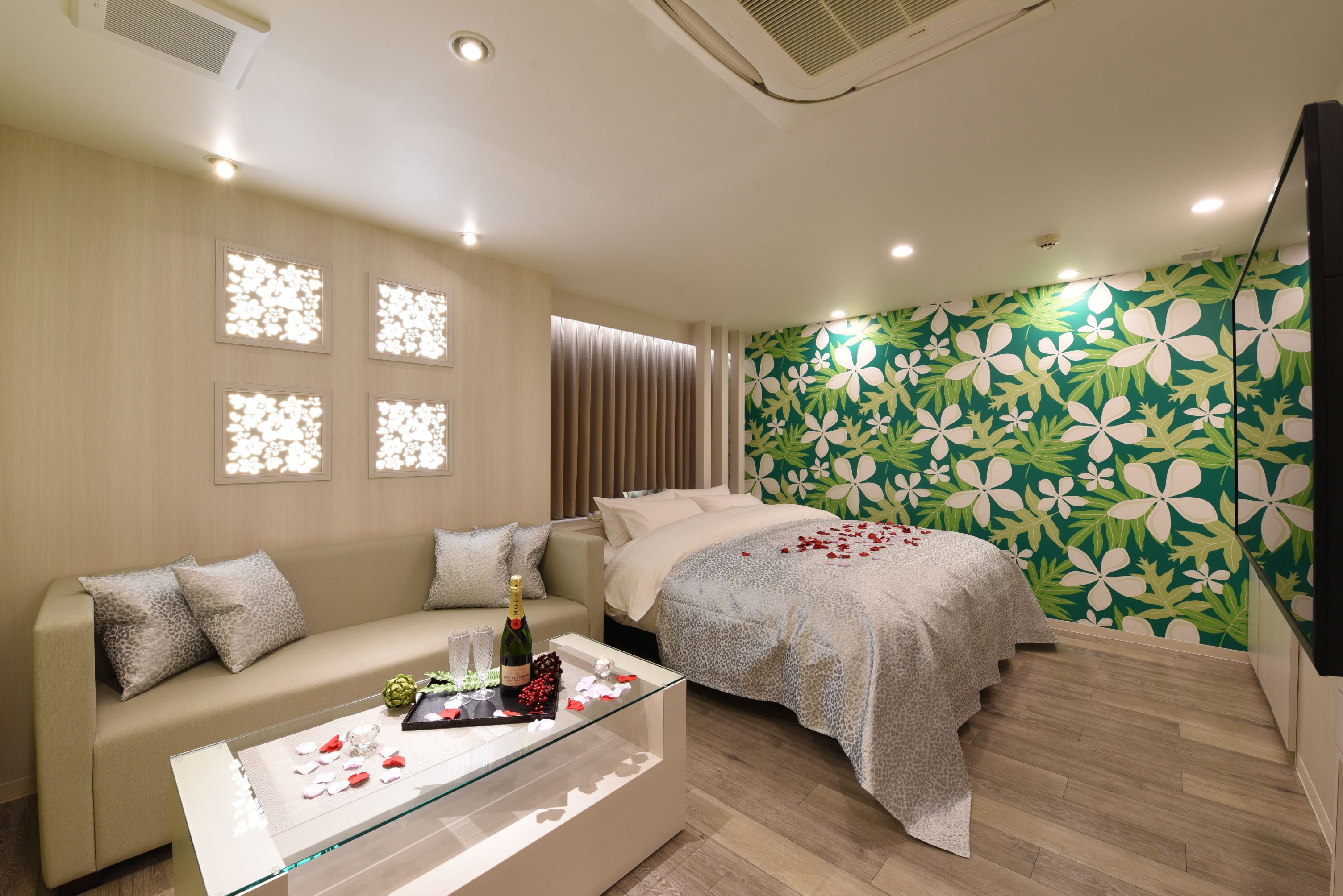 A modern bedroom with a sofa, a bed, and a glass coffee table with a bottle and flowers.